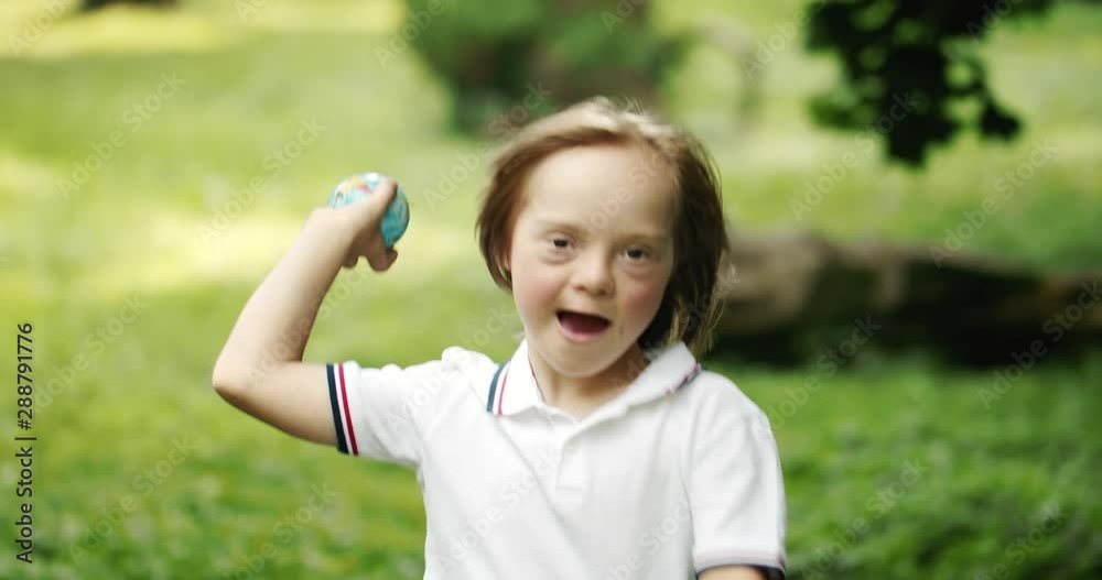 Little boy with Down Syndrome playing ball in green lawn in park, running with mother Little boy with Down Syndrome playing ball in green lawn in park, running with mother