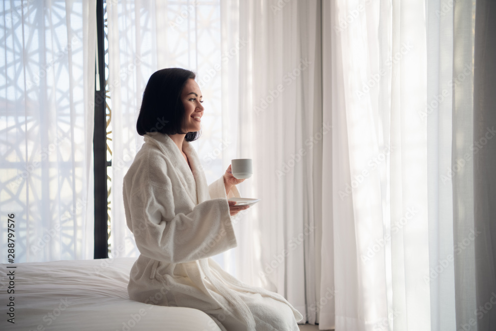 Attractive young girl in bathrobe holding a cup, looking at window and smiling while sitting on a bed near the big window at home Attractive young girl in bathrobe holding a cup, looking at window and smiling while sitting on a bed near the big window at home