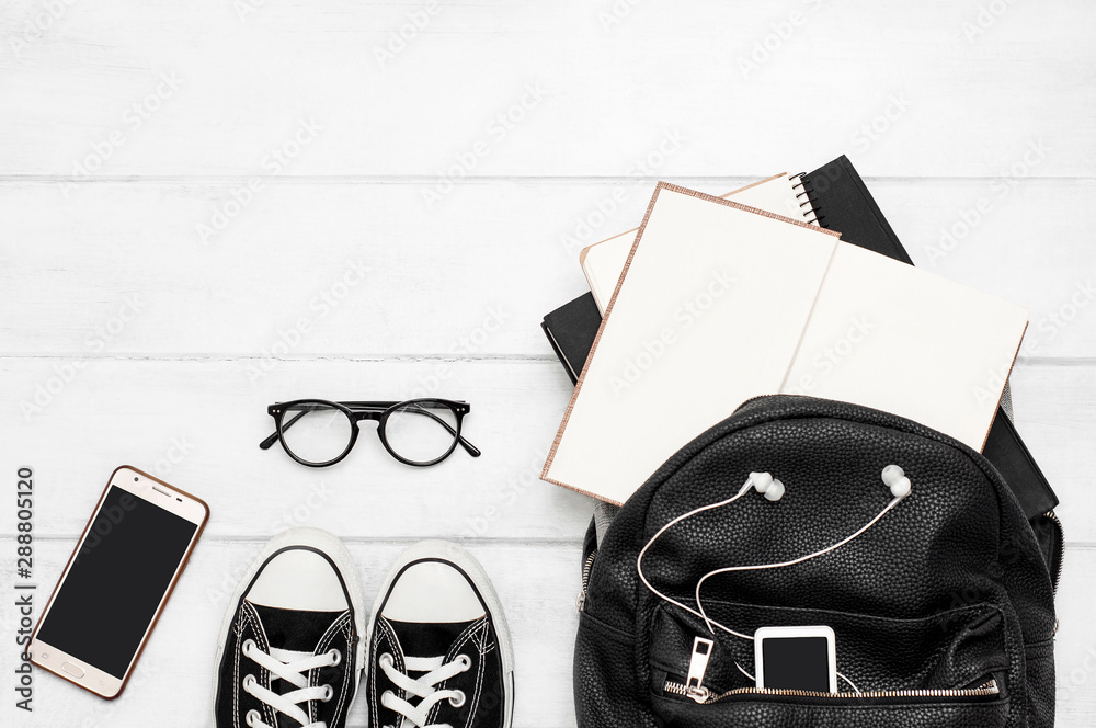 An open black backpack with books, a mobile phone, a player with headphones, glasses, notebook, gym shoes on a white wooden background. Back to school. Student. Top view. Flat lay - obrazy, fototapety, plakaty 