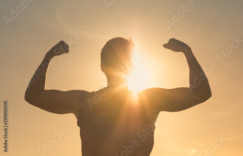 Strong fit man flexing against sunset sky. People power and winning concept. 