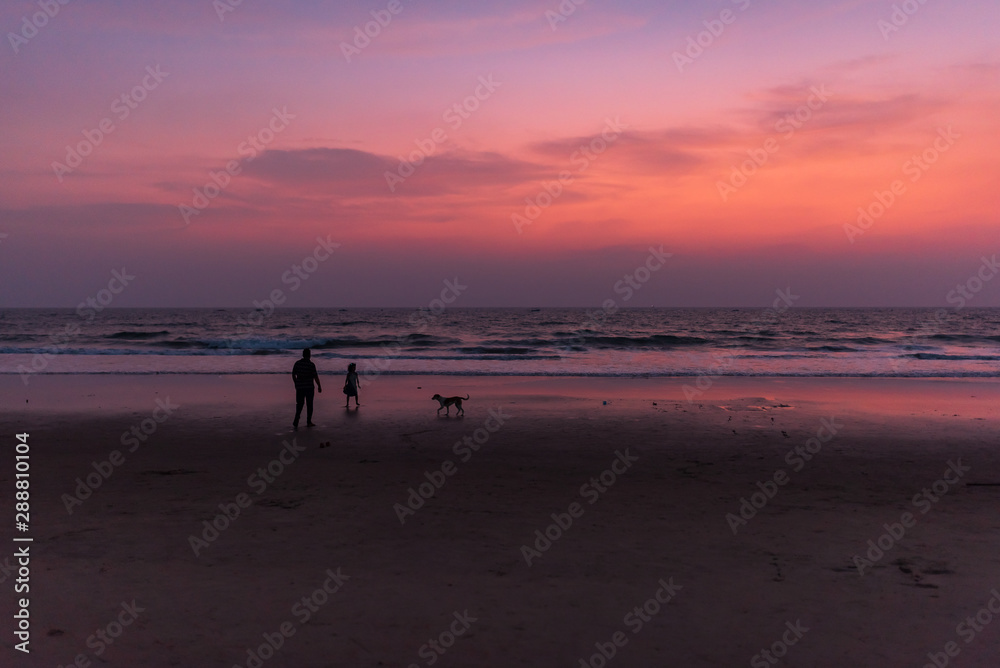 sunset at a beach, with yellow, pink and orange sky. Father, daughter and pet dog playing at the beach.
