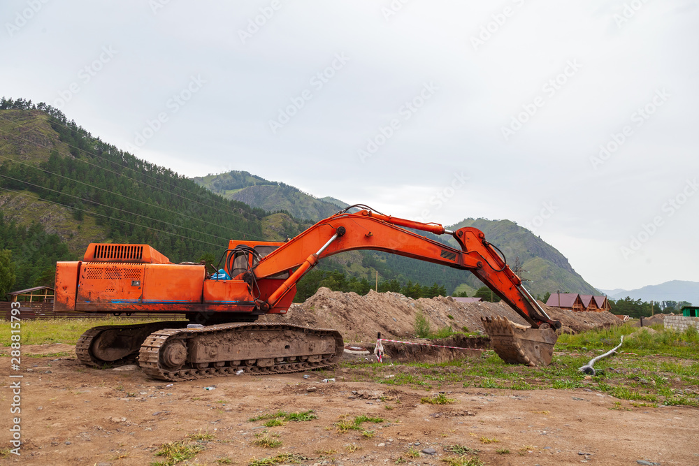 Large orange excavator with a lowered bucket during repair work in the mountains on a summer day. Large orange excavator with a lowered bucket during repair work in the mountains on a summer day.