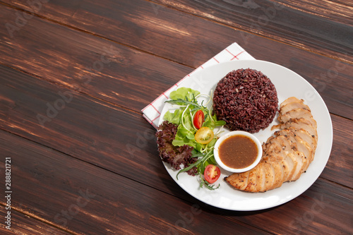 Top view of roasted chicken breast fillet steak with brown rice/coarse rice, vegetables salad and spicy sauce in white plate on wooden table. Healthy food menu, eating right concept.