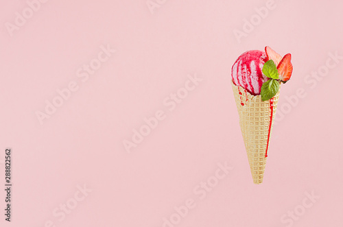 Pink ice cream in crisp waffle cone with strawberry slices, red sweet sauce, green mint leaf on pink background.
