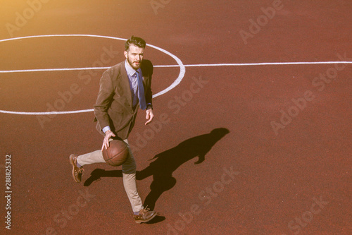An attractive business guy in a suit and tie runs along a sports basketball court with a ball in his hands. Active lifestyle and sports office worker. Hipster plays basketball. Bearded stylish man. © Daniil