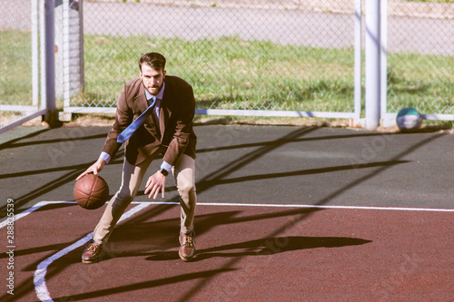 Stylish bearded handsome hipster guy in a trendy suit in a jacket and tie on a basketball court playing basketball. Fashionable handsome man with a ball in his hand, top view. Business concept, sport.