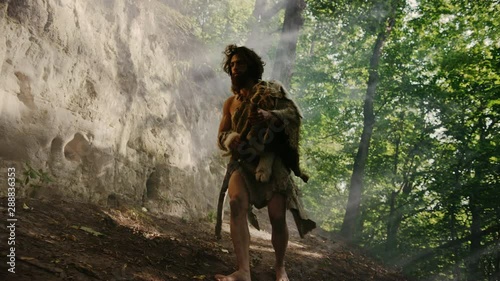 Primeval Caveman Wearing Animal Skin Holds Stone Tipped Hammer Comes out of the Cave and Looks Around, Exploring Prehistoric Forest Ready to Hunt Animal Prey. Neanderthal Going Hunting into the Jungle