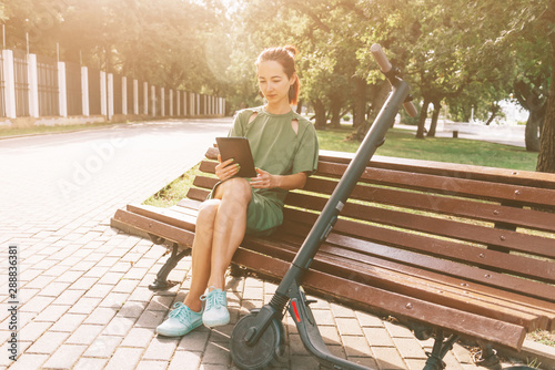 Girl resting on bench with digital tablet and an e-scooter in park.