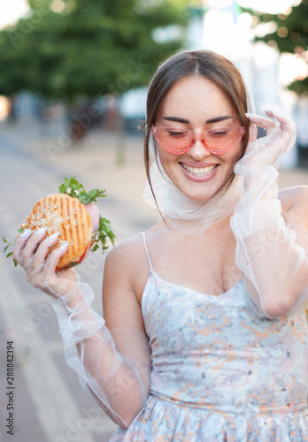 Charming girl in pink sunglasses and a dress eats a hamburger on the street