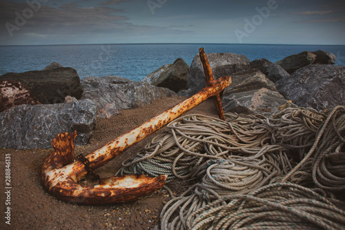 A rusty anchor and rope set on the rocks with the English channel sea in the background.