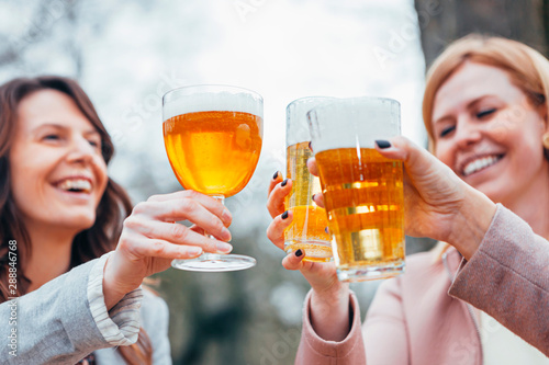 three women sitting at an outdoor terrace in autumn, talking, laughing and toasting together with beer.