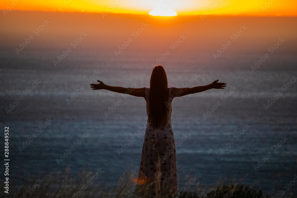Fototapeta premium Silhouette of a beautiful woman standing with her arms spread open at sunset or sunrise.