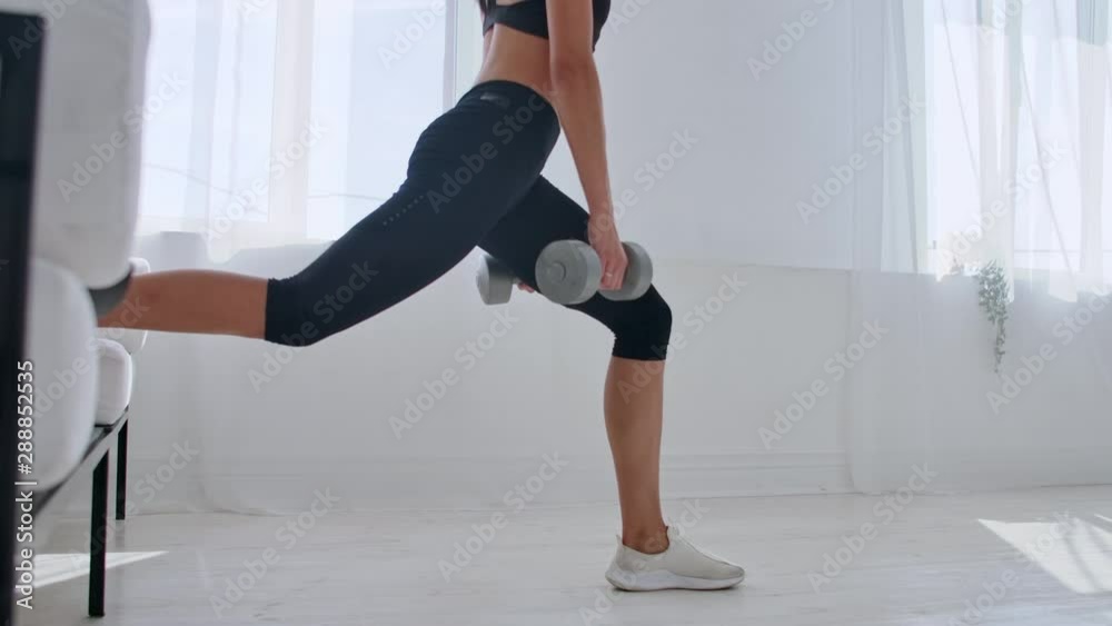 Brunette in black sportswear in a white apartment makes a split squat with dumbbells in her hands leaning on the sofa with her foot Brunette in black sportswear in a white apartment makes a split squat with dumbbells in her hands leaning on the sofa with her foot