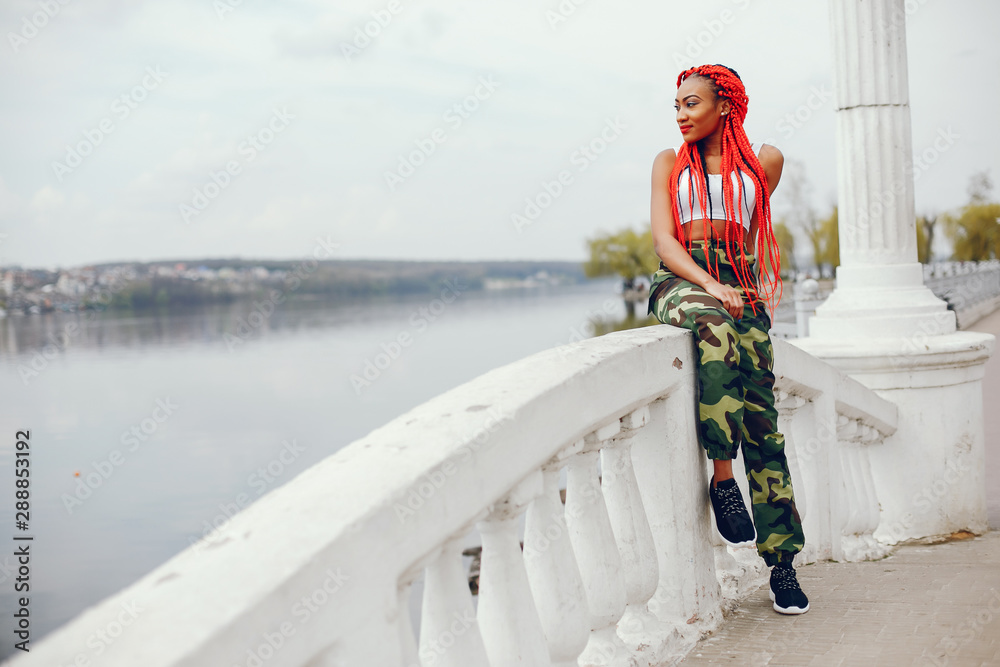 A young and stylish dark-skinned girl with red dreads walking in the summer park near river
