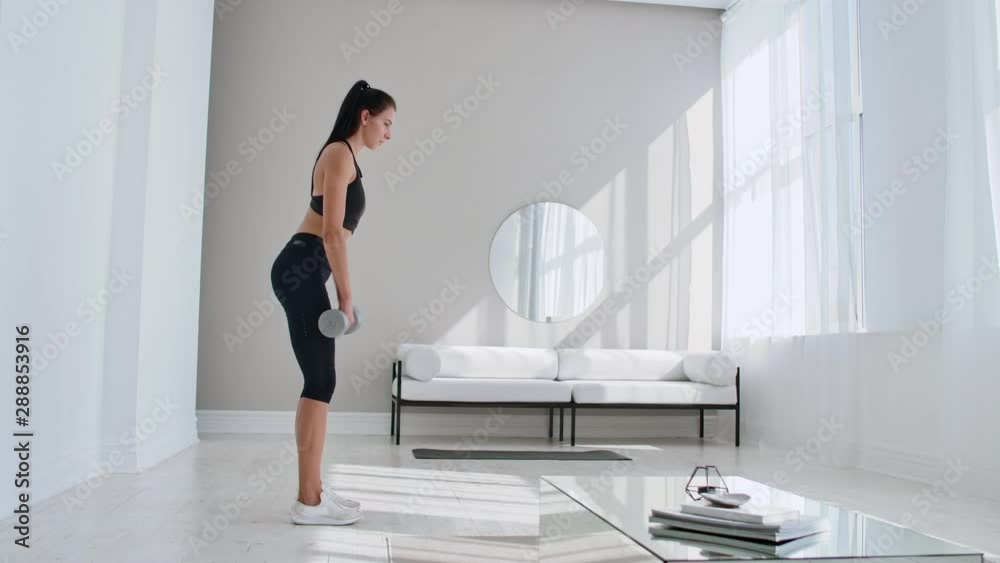 The beautiful female performs leans forward with dumbbells in her hands in the white interior of the living room. In slow motion to do exercises on the muscles of the thighs.