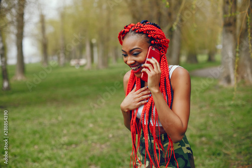 A young and stylish dark-skinned girl with red dreads walking in the summer park with phone