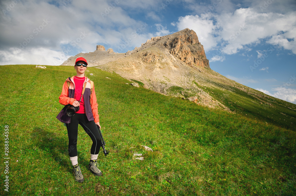 Girl athlete photographer in a red cap sunglasses and a yellow backpack stands on a green slope against the background of the epic cliffs of the Caucasus