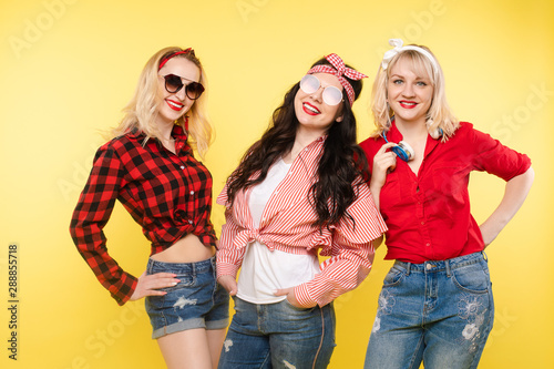 Studio shot of trendy girls with head bows, sunglasses wearing trendy hipster clothes and smiling at camera. Isolate on yellow background.