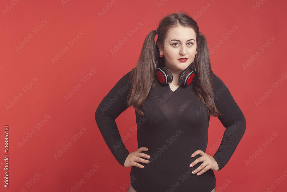 Model plus size with sweet donut, happy girl posing with headphones, in black bodysuit. XXL female on red background Model plus size with sweet donut, happy girl posing with headphones, in black bodysuit. XXL female on red background