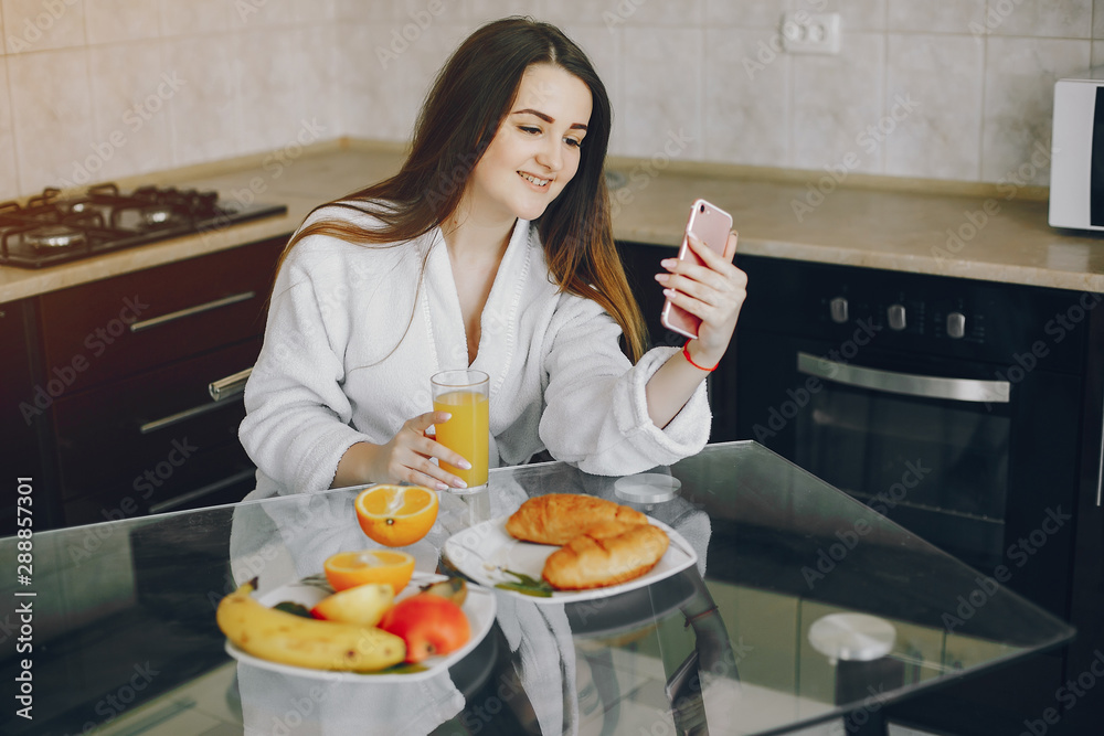 beautiful young girl with black hair and white robe sitting at home in the kitchen at the table and eating an croissant and drinking orange juice