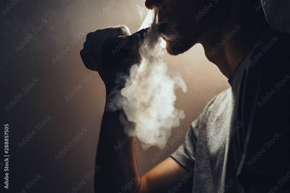 Closeup of a vaping man holding a mod. A cloud of vapor. Black background.