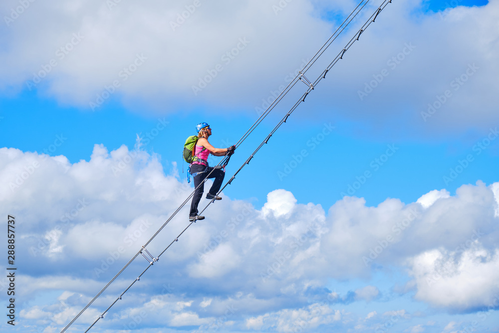 Stairway to heaven concept. Woman climbing a diagonal via ferrata ladder in Donnerkogel mountains, Austria, near Gosau, with white clouds and blue sky behind her.