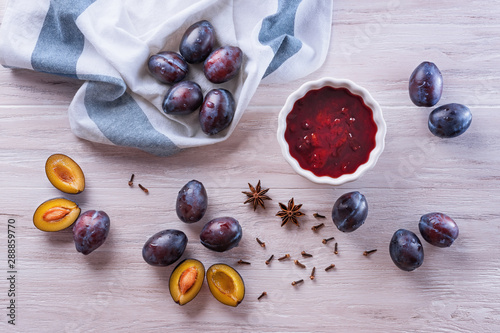 Fresh ripe plums, a white ceramic bowl with homemade plum jam, creasy towel, badyan seeds and dry carnation grains on a wooden table. Flat lay, top view photo