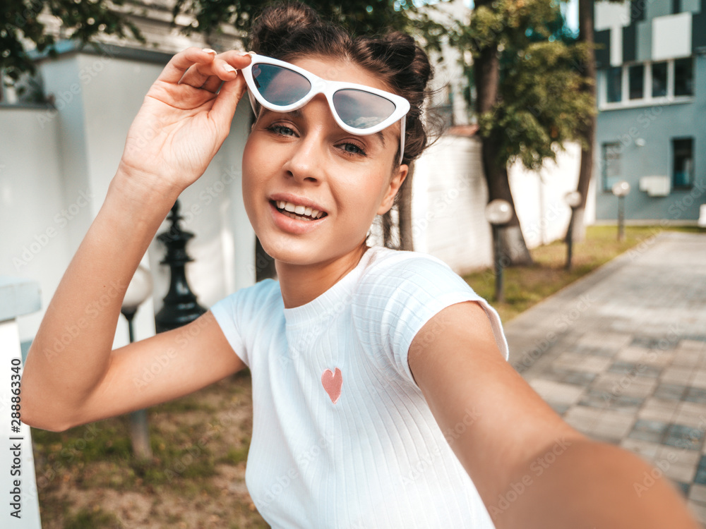 Beautiful smiling model with horns hairstyle dressed in summer casual clothes.Sexy carefree girl posing in the street in sunglasses.Taking selfie self portrait photos on smartphone Beautiful smiling model with horns hairstyle dressed in summer casual clothes.Sexy carefree girl posing in the street in sunglasses.Taking selfie self portrait photos on smartphone