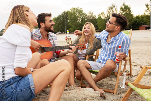 Group of friends sitting at the beach and play guitar and singing.