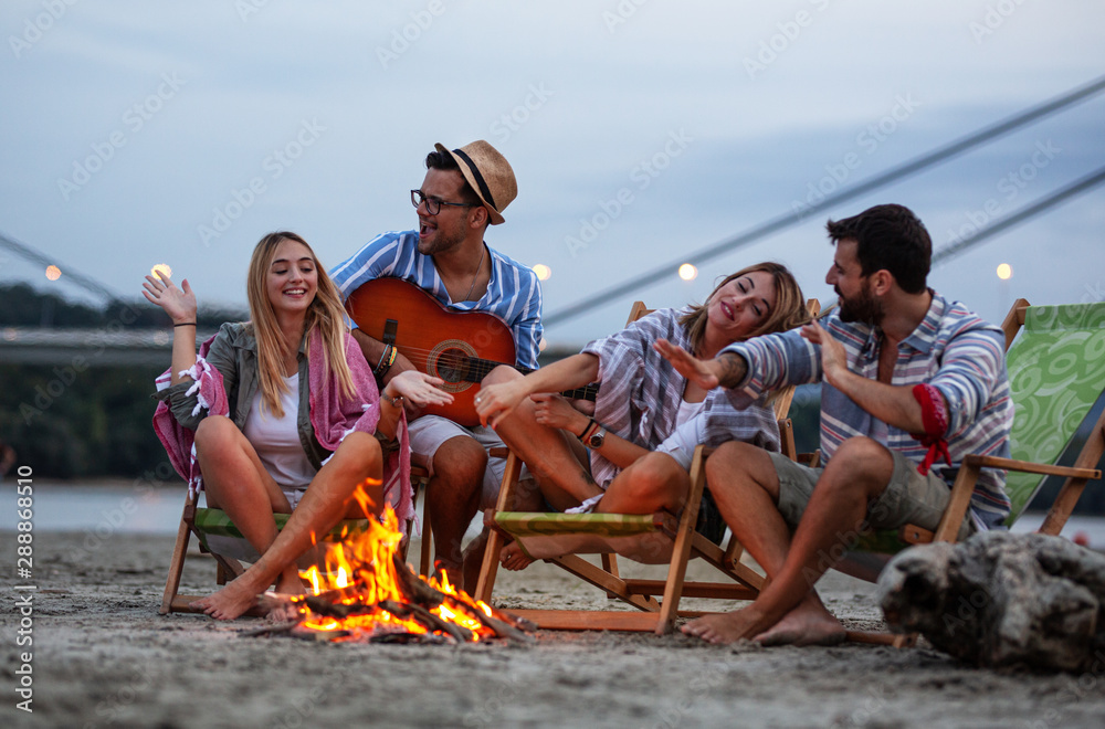 Obraz premium Group of friends sitting around camp fire at the beach at the evening.They play guitar and singing.