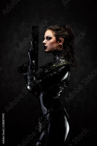 Portrait of a young woman in latex overalls holding a futuristic weapon in her hands