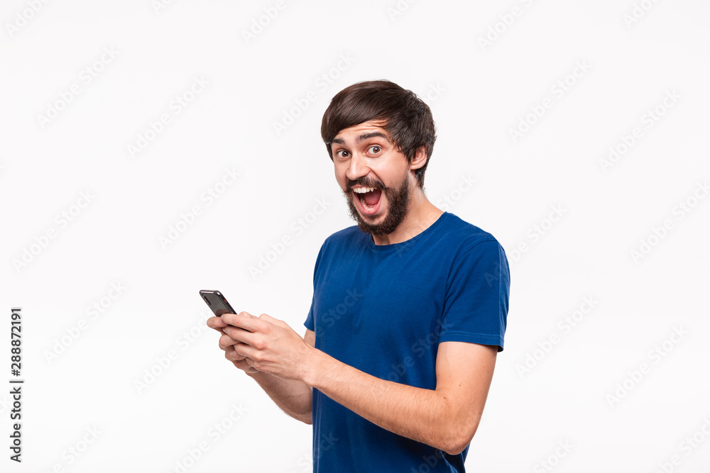 Handsome brunet man in a blue shirt with beard and mostaches surprised holding smartphone looking at the camera standing isolated over white background. Handsome brunet man in a blue shirt with beard and mostaches surprised holding smartphone looking at the camera standing isolated over white background.