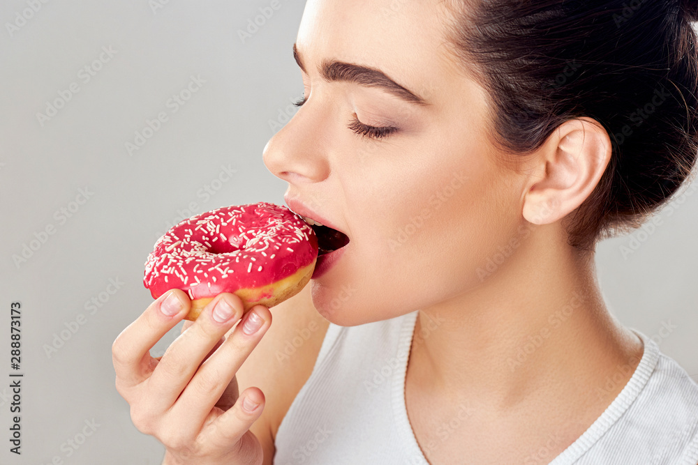 Close up portrait of a hungry girl eating donuts isolated over white background.Brunette girl holds and tastes a pink donut. Sweets are unhealthy junk food. Dieting. Close up portrait of a hungry girl eating donuts isolated over white background.Brunette girl holds and tastes a pink donut. Sweets are unhealthy junk food. Dieting.