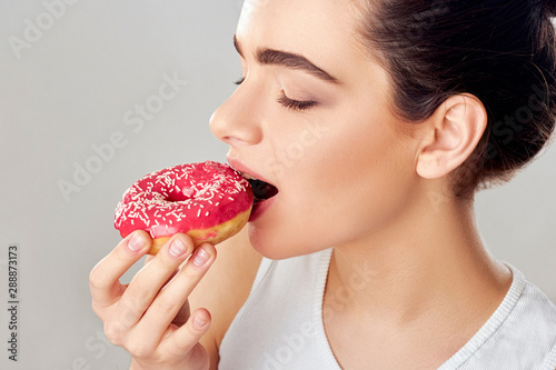 Close up portrait of a hungry girl eating donuts isolated over white background.Brunette girl holds and tastes a pink donut. Sweets are unhealthy junk food. Dieting. 