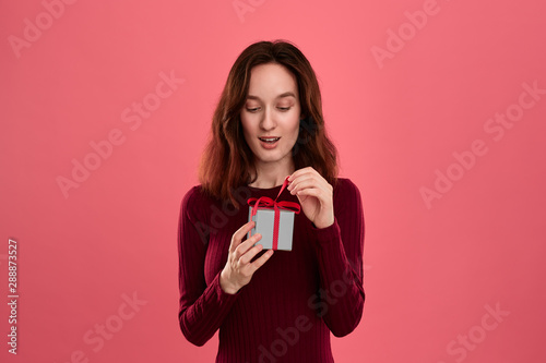 Excited pretty brunette girl opens gift packed in present box with a ribbon standing isolated on a dark pink background and smiling at the camera. Celebrating special event. © ianachyrva