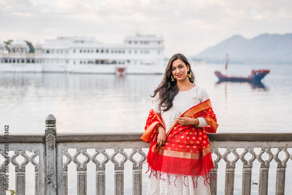 Indian female model at Ambrai Ghat in front of Taj Palace in Udaipur, Rajasthan, India. Hindu girl with traditional Indian white dress with gold ornaments and red wedding dupatta. 