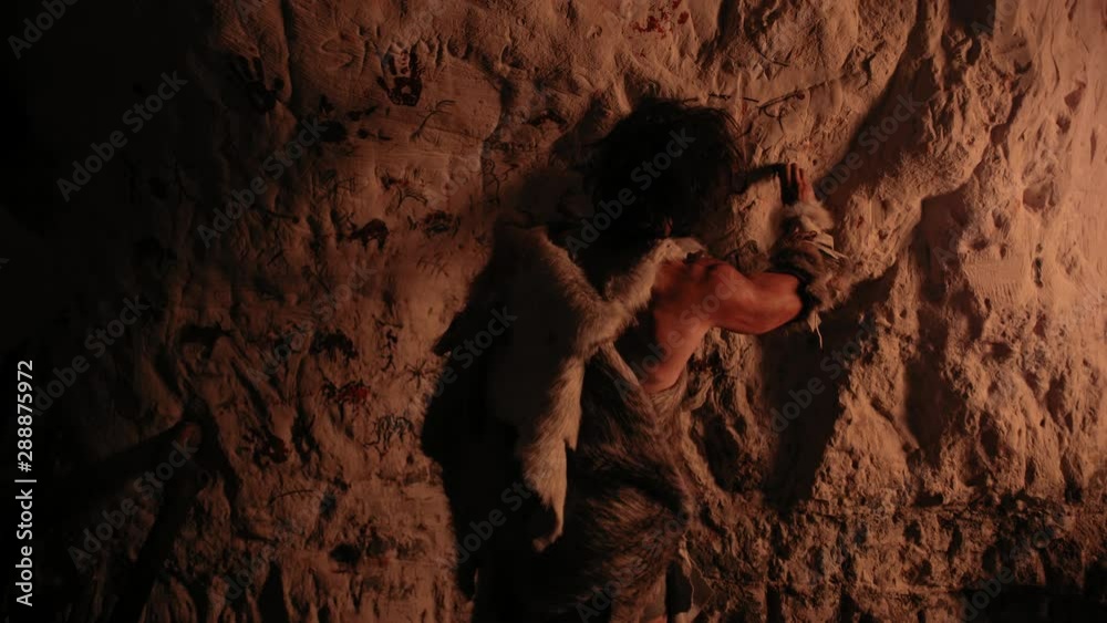 Primitive Prehistoric Neanderthal Wearing Animal Skin Draws Animals and Abstracts on the Walls at Night. Creating First Cave Art with Petroglyphs, Rock Paintings Illuminated by Fire. Back View Zoom In Primitive Prehistoric Neanderthal Wearing Animal Skin Draws Animals and Abstracts on the Walls at Night. Creating First Cave Art with Petroglyphs, Rock Paintings Illuminated by Fire. Back View Zoom In