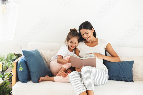 Image of attractive family pregnant woman and her little daughter reading book while sitting on sofa at home