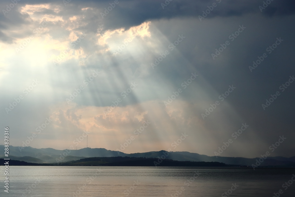Dark sea and cloudy sky with sun rays lightning through the clouds. Green hills and mountains on the horizon. Landscape and seascape for design template, background or wallpaper.