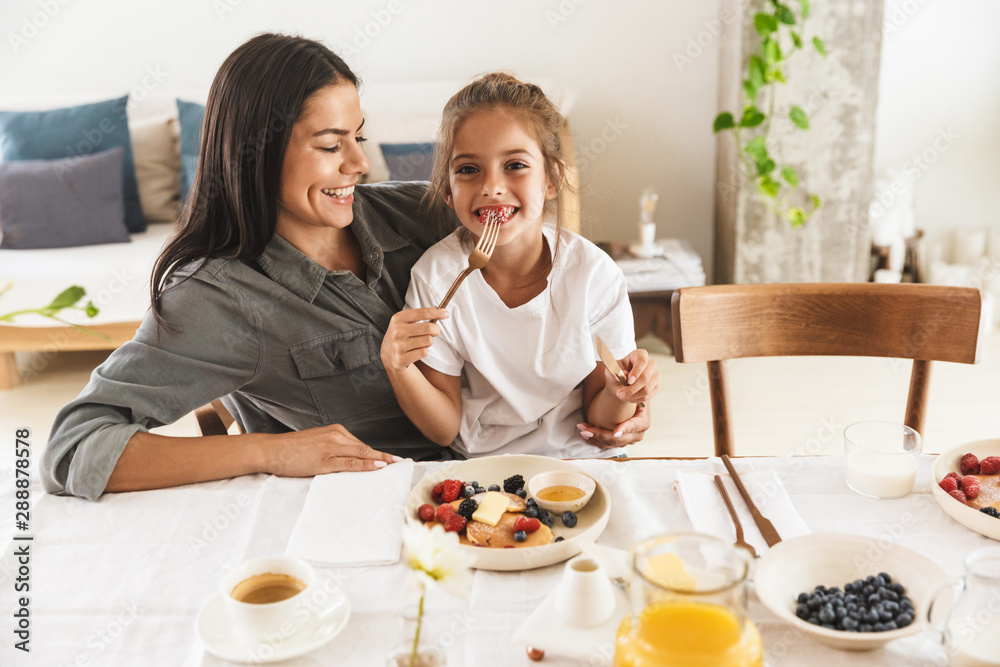 Image of joyous family mother and little daughter smiling and eating together while having breakfast at home in morning