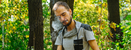 A young handsome man with beard in casual clothes is smoking a cigarette with closed eyes in the forest.