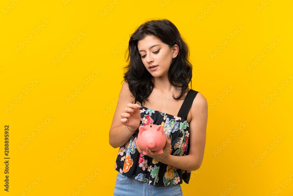 Young woman over isolated yellow background holding a big piggybank Young woman over isolated yellow background holding a big piggybank