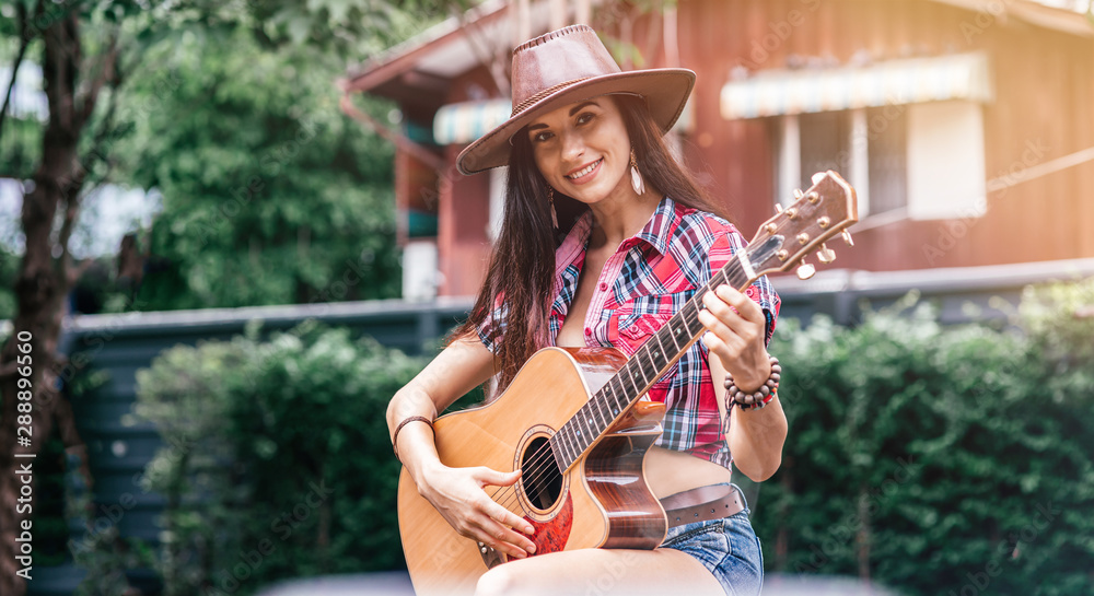 Fototapeta premium Portrait of caucasian young woman in checkered shirt and cowboy hat. She is looking at camera with a smile while playing country music using guitar outdoors. Living on a farm concept. Horizontal shot