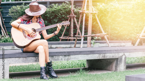Portrait of caucasian young woman in cowgirl outfit. She is playing country music using her guitar while sitting outdoors. Living on a farm concept. Horizontal shot