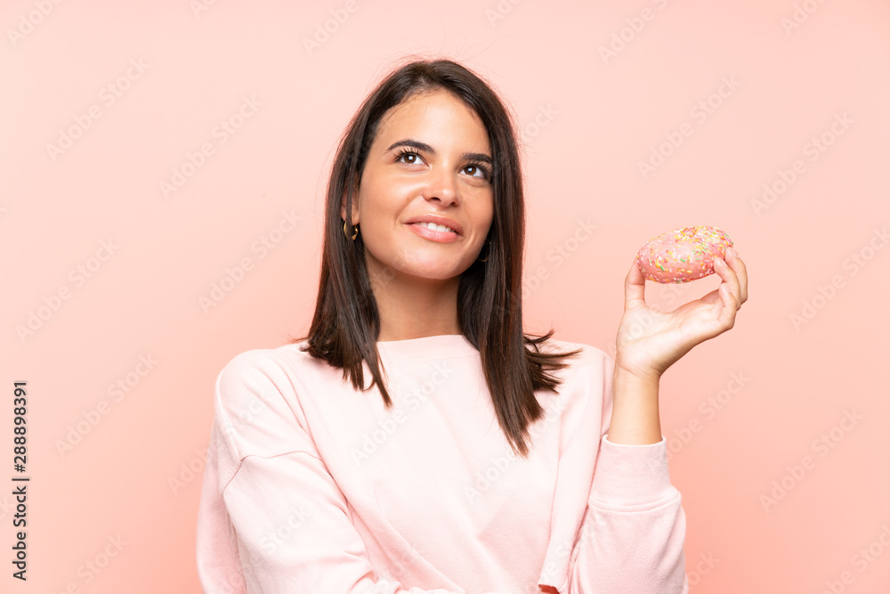 Young girl holding a donut over isolated pink background looking up while smiling Young girl holding a donut over isolated pink background looking up while smiling
