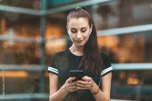 Portrait of young woman looking at the screen of her phone with a smile while standing outdoors and listening to music or podcast via wireless earphones. Horizontal shot. Selective focus. Front view