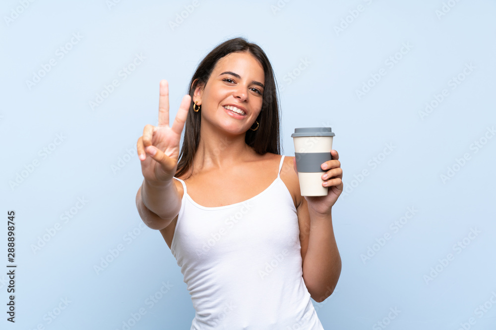 Young woman holding a take away coffee over isolated blue background smiling and showing victory sign Young woman holding a take away coffee over isolated blue background smiling and showing victory sign