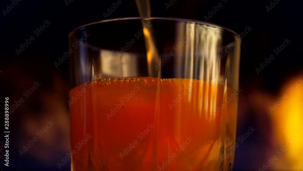 A yellow-orange sweet multifruit juice is poured into a glass with ice cubes. Glass with juice on a background of colored smoke. A yellow-orange sweet multifruit juice is poured into a glass with ice cubes. Glass with juice on a background of colored smoke.