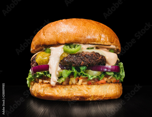Craft burger is cooking on black background. Consist: sauce salsa, lettuce, red onion, pickle, cheese, chilli green pepper, bun, marbled meat beef.