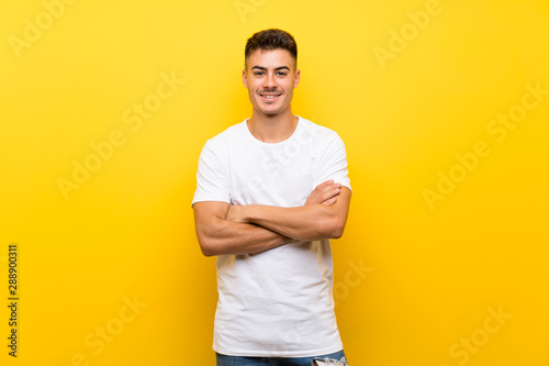 Young handsome man over isolated yellow background keeping the arms crossed in frontal position